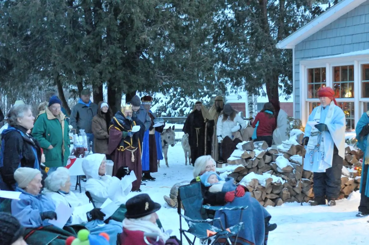 People gather in the snow for an outdoor pageant beside a house and a large stack of firewood.