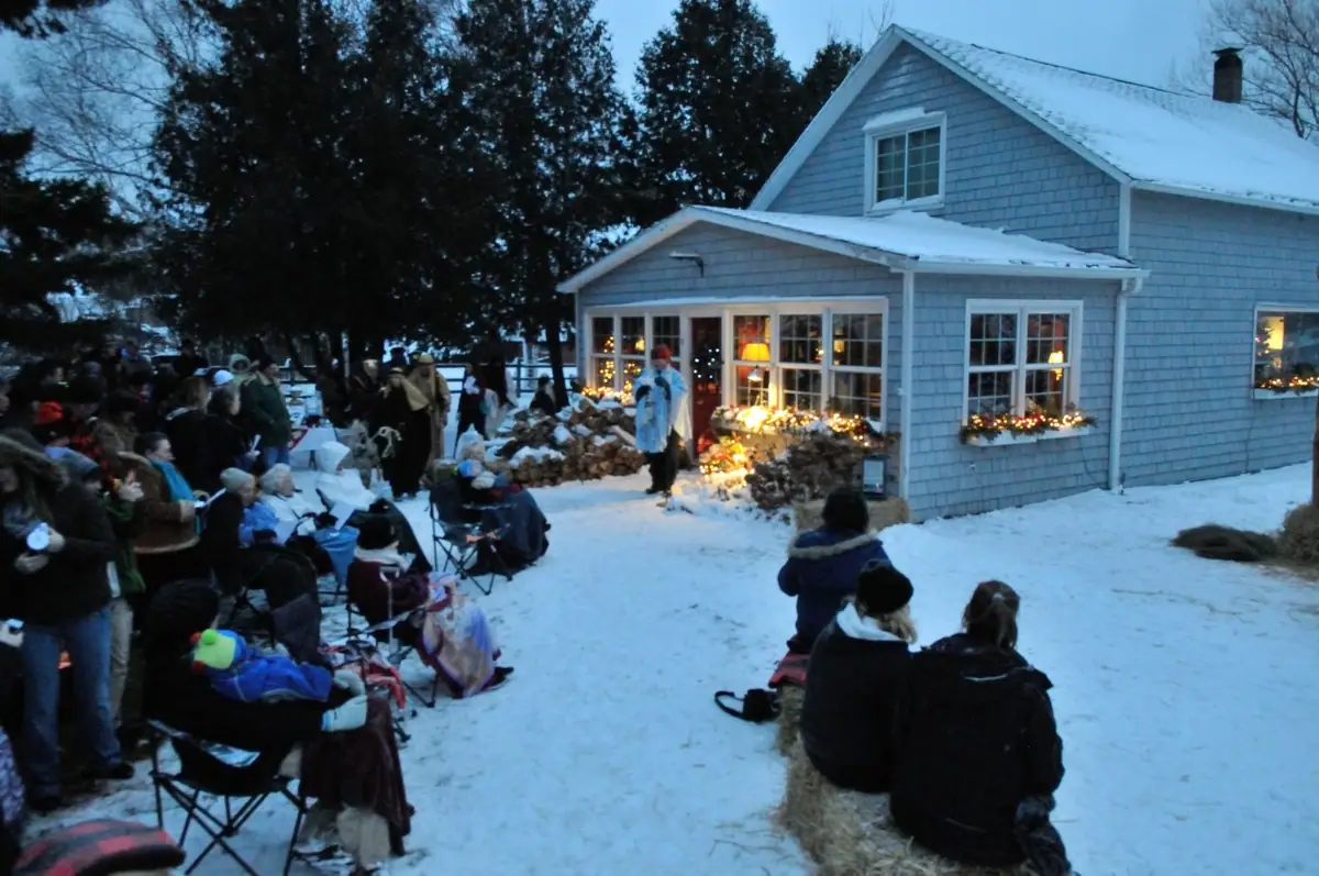 A winter nativity performance takes place outside a light-blue house as people watch from chairs and hay bales.