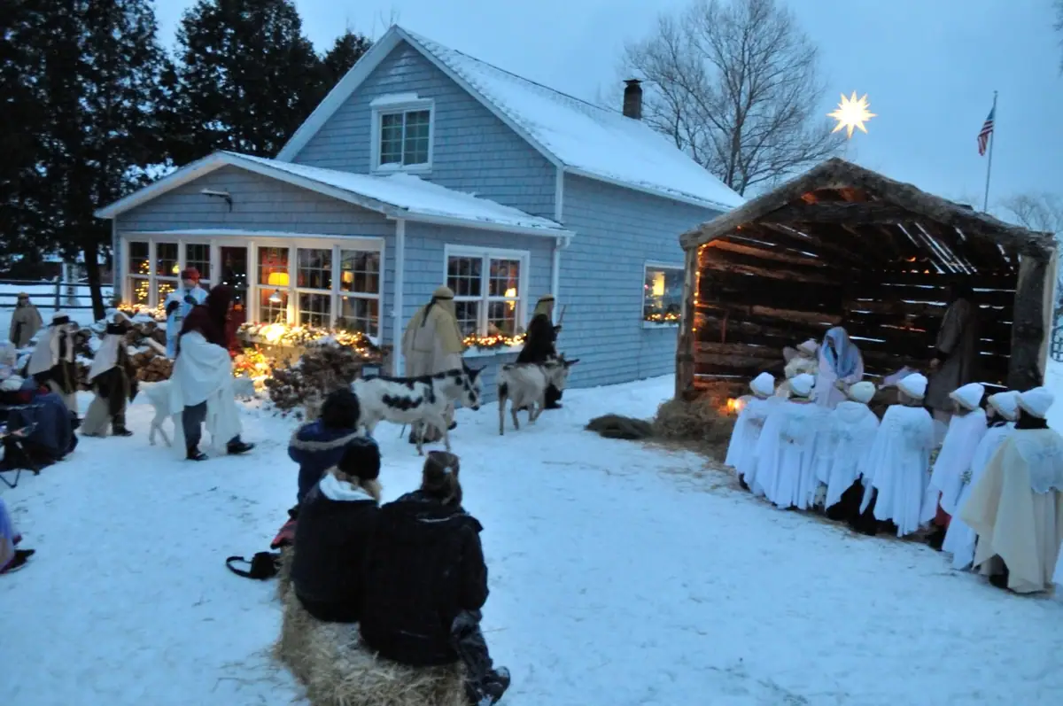 A snow-covered outdoor tableau with robed participants, goats, a rustic shelter, and a lighted star beside a house.