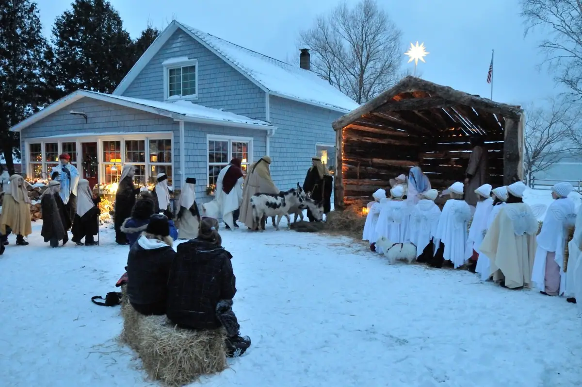 A costumed nativity scene unfolds in snow beside a blue house and a rustic stable.