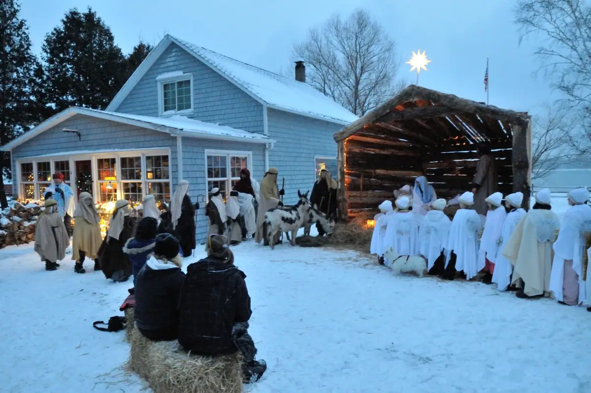 Costumed participants gather in a snowy yard beside a rustic shelter, with two donkeys and seated spectators on a hay bale.