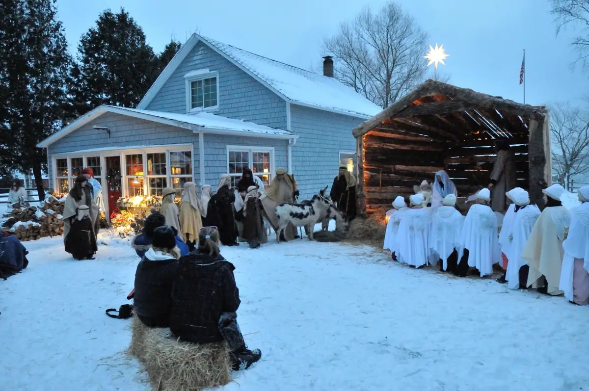 People in robes gather around a rustic shelter beside a house during a snowy outdoor nativity scene.