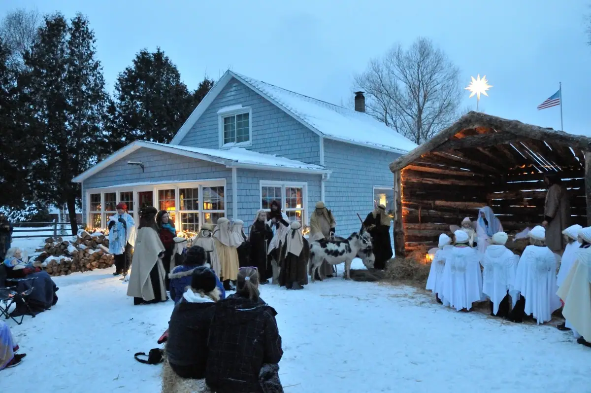 Outdoor winter nativity scene with costumed participants, goats, a stable, and seated children.