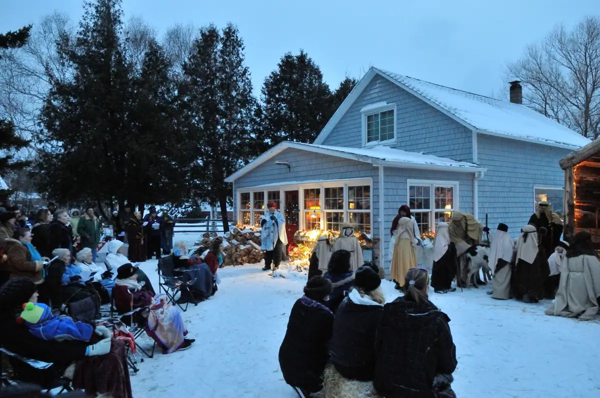 A costumed nativity scene unfolds outside a snow-covered house as an audience watches.
