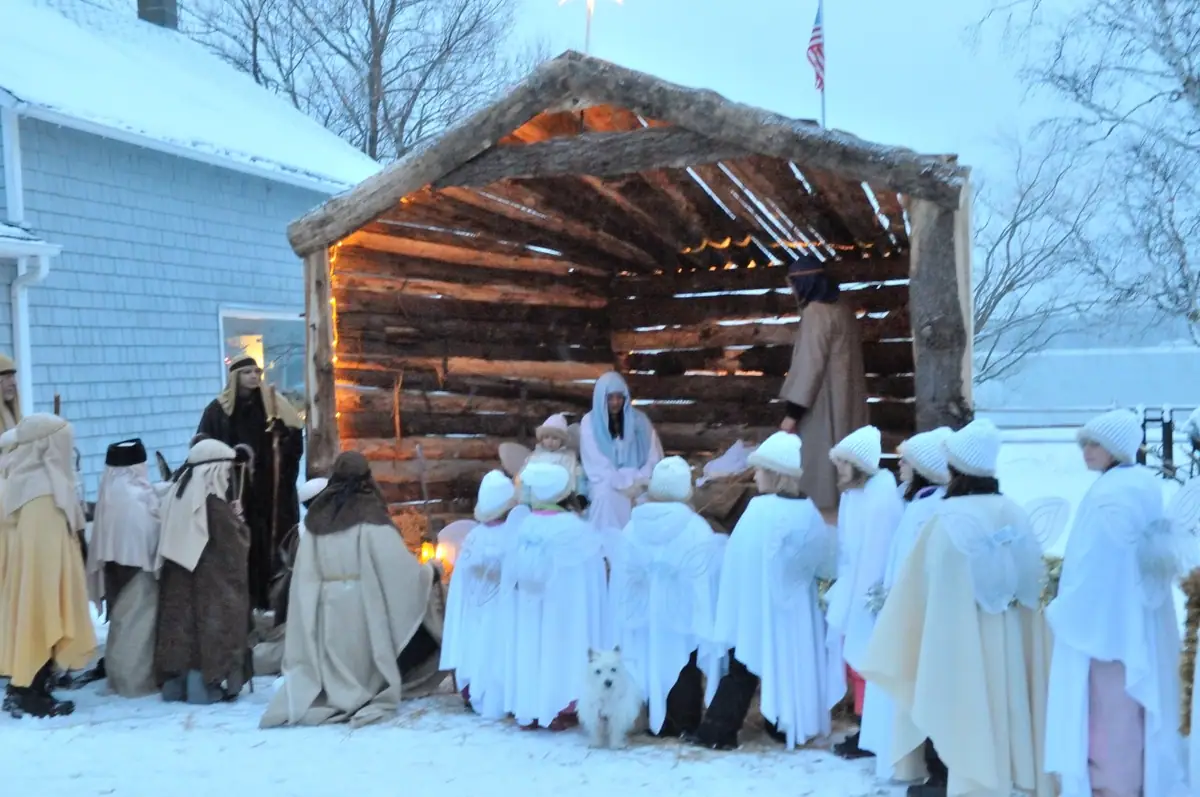 Costumed participants gather around a log shelter for an outdoor nativity scene in the snow.