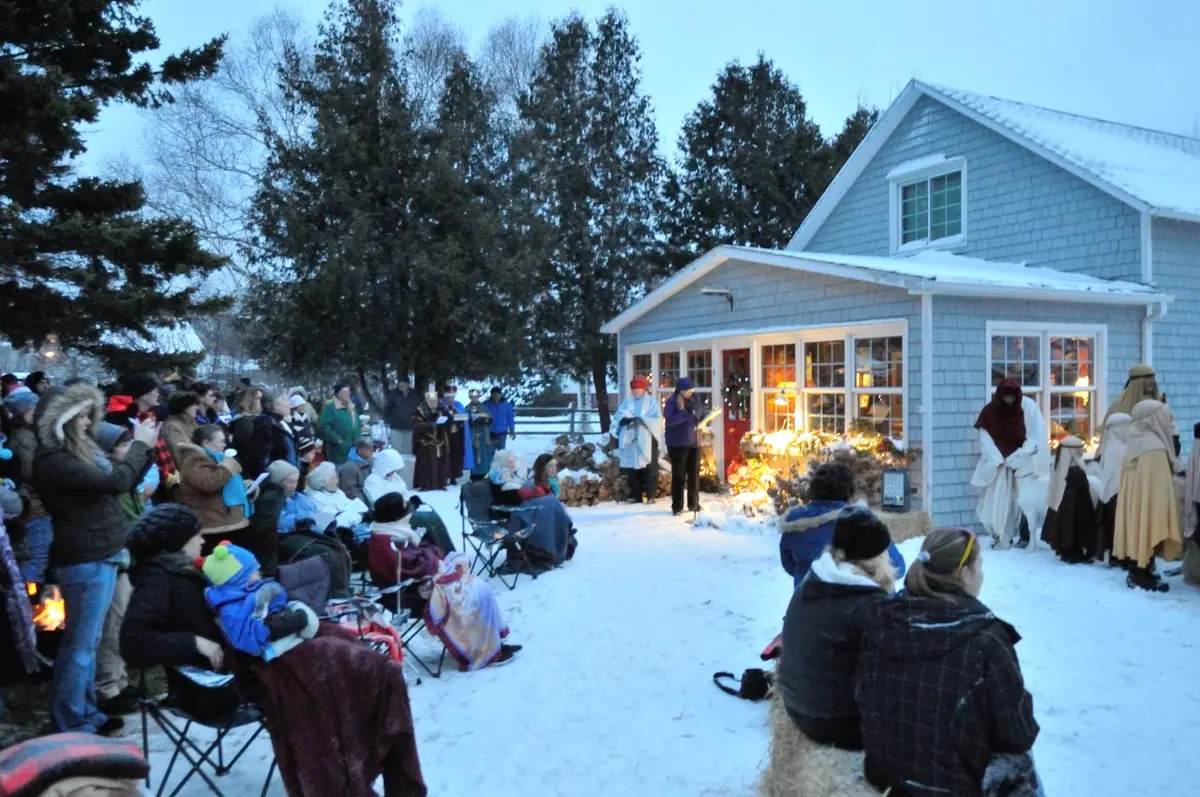 A winter outdoor gathering with robed figures, seated spectators, and a snow-covered shingled house.