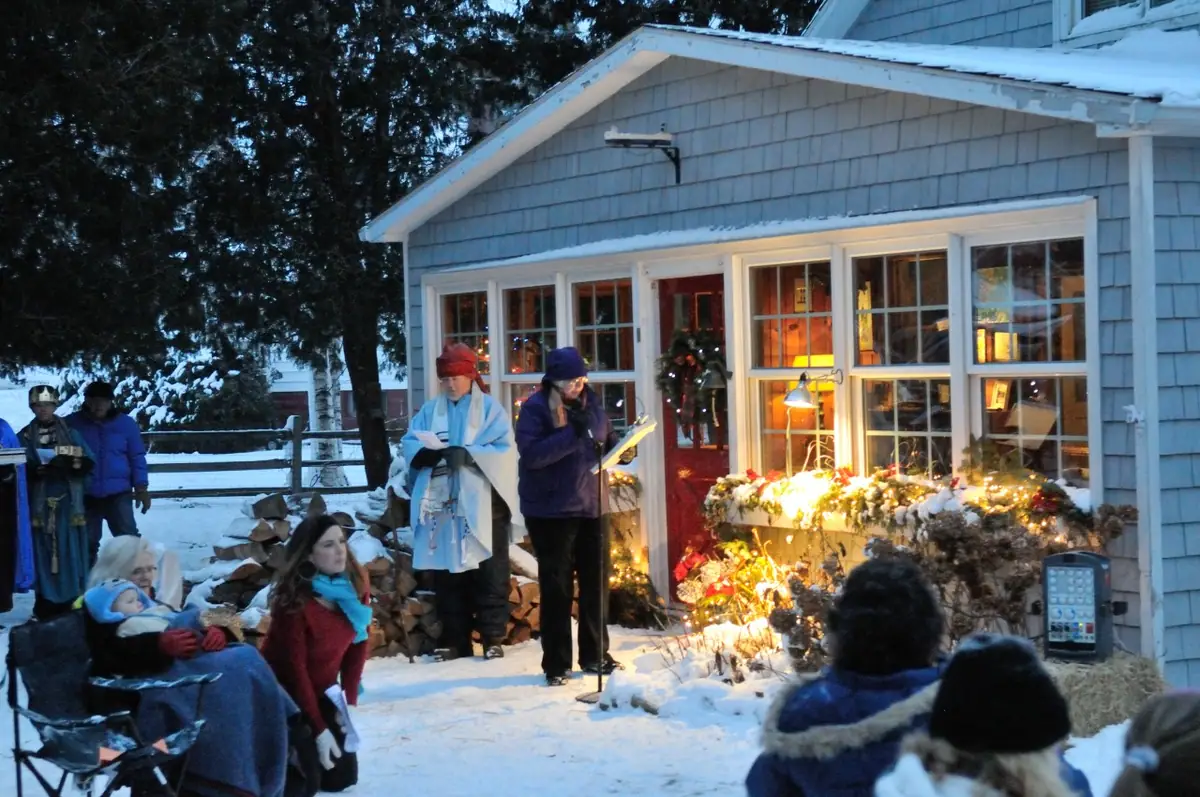 People gather in the snow outside a shingled building while a participant reads from a paper.