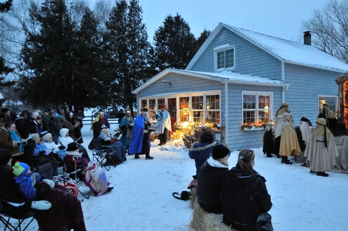 People in winter clothing and robes gather in a snowy yard beside a decorated house.