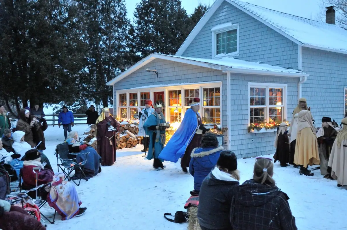 People in biblical costumes perform outdoors in the snow beside a decorated shingled house as spectators watch.