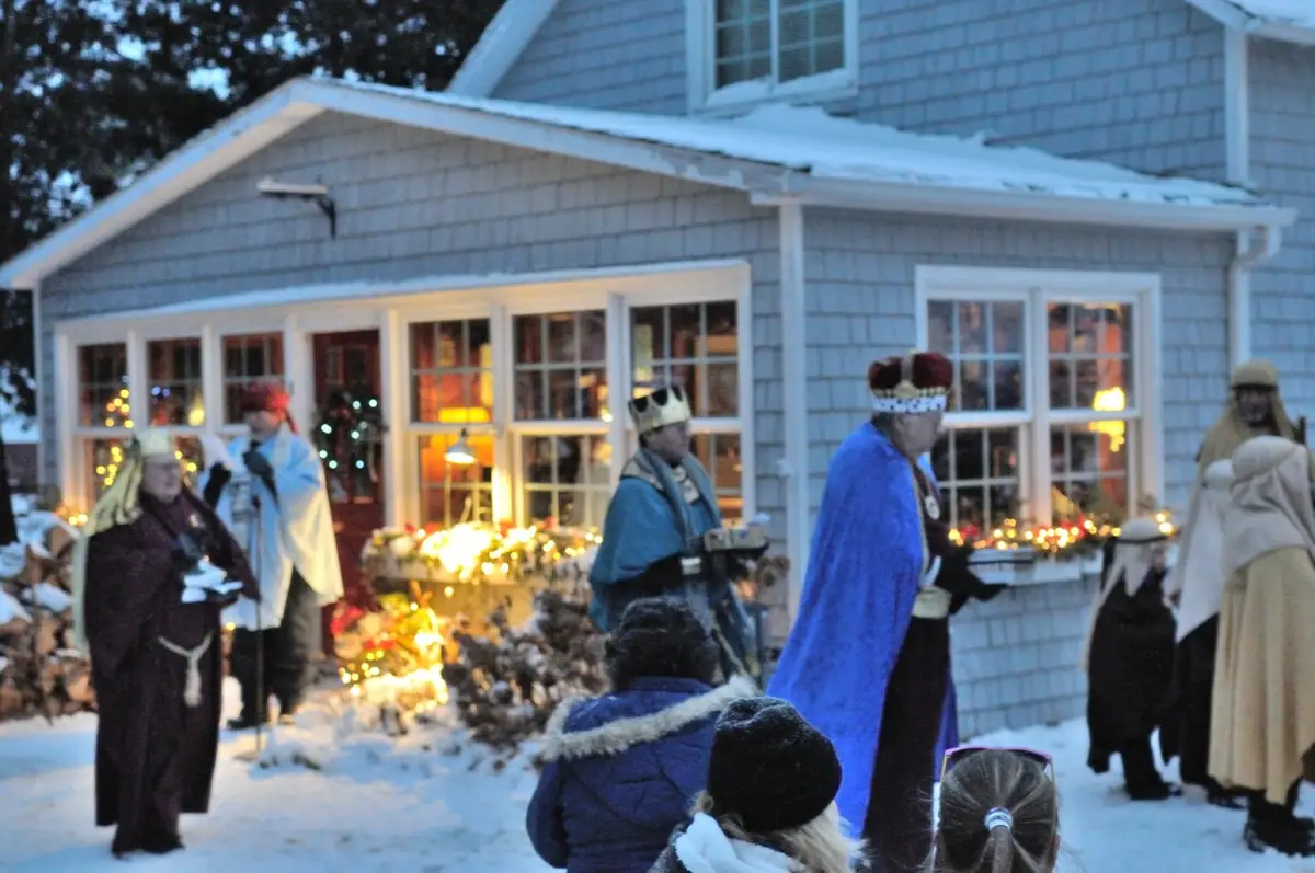 Costumed people gather in the snow outside a shingled house as children watch a nativity scene.