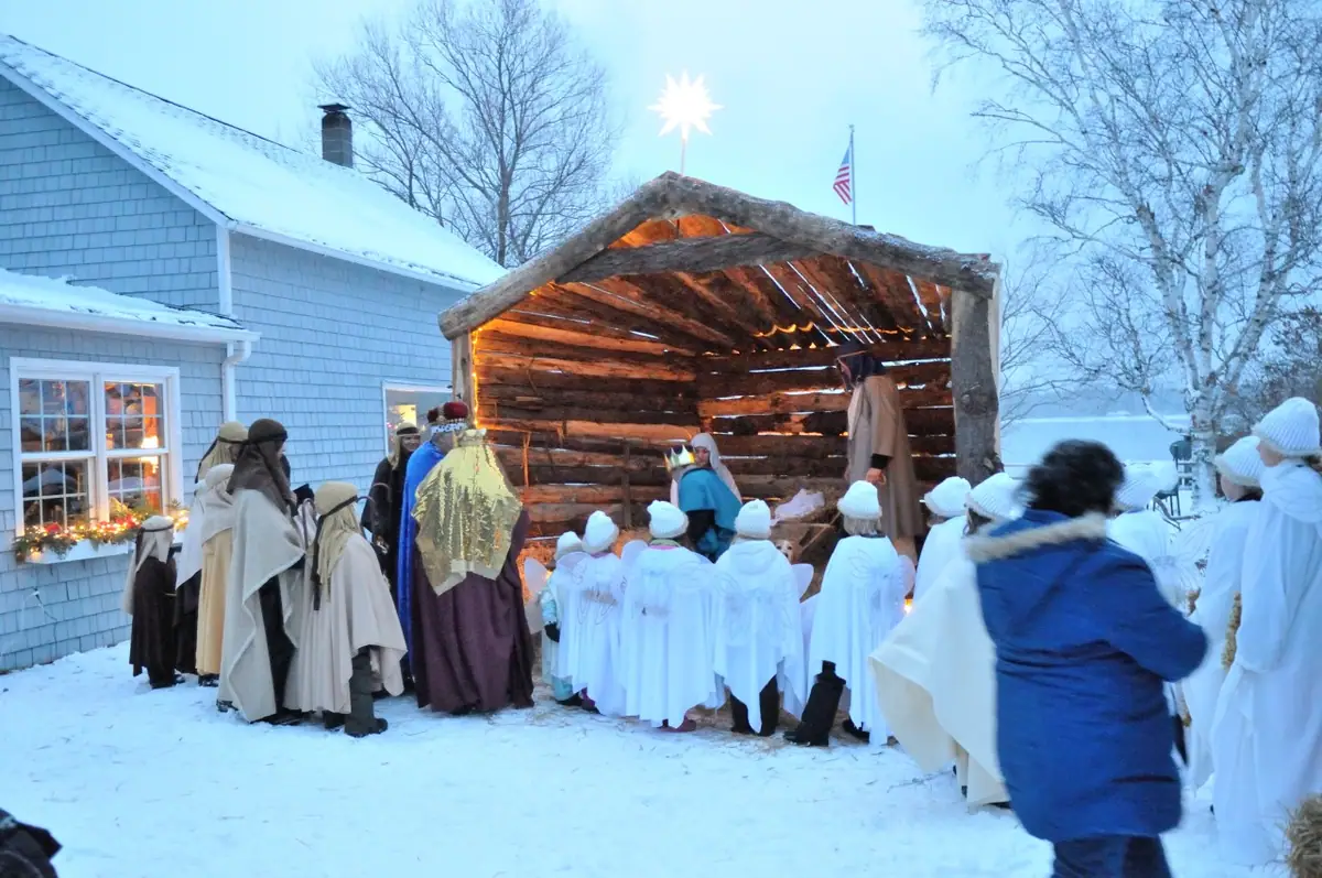 Costumed performers and children gather around a lit wooden nativity shelter in a snowy yard.