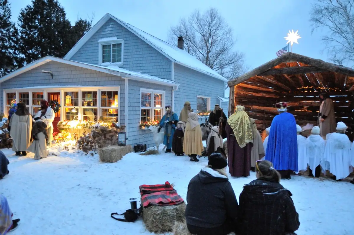 A costumed nativity scene unfolds in a snowy yard beside a lit house and rustic stable.
