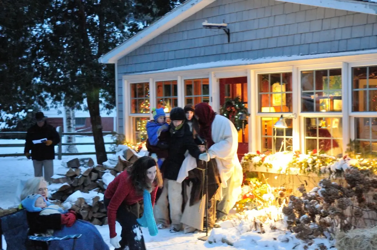 Adults and children in winter clothing gather outside a decorated, snow-covered house at dusk.