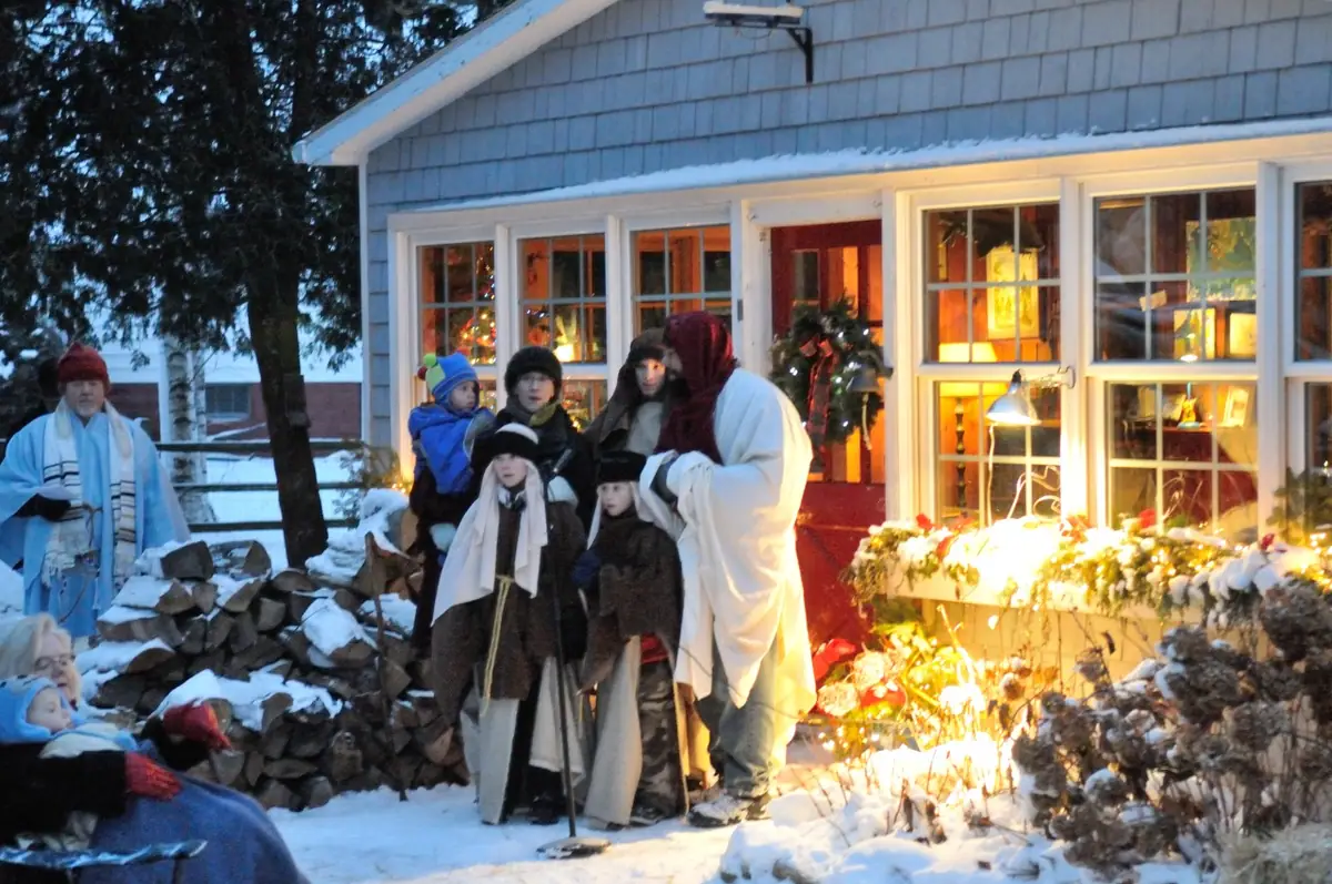 People in robes and winter clothing gather in the snow outside a brightly lit house.