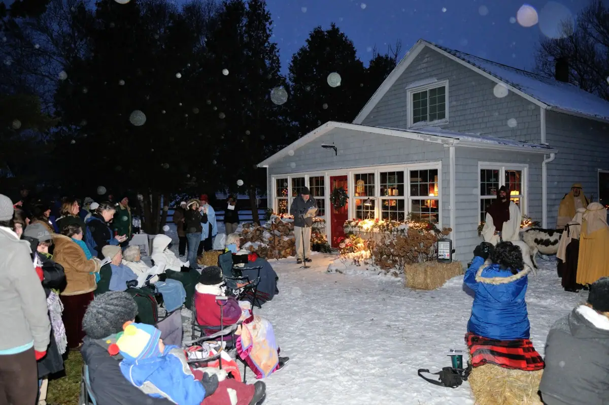 A crowd watches a costumed outdoor tableau beside a brightly lit house on a snowy night.