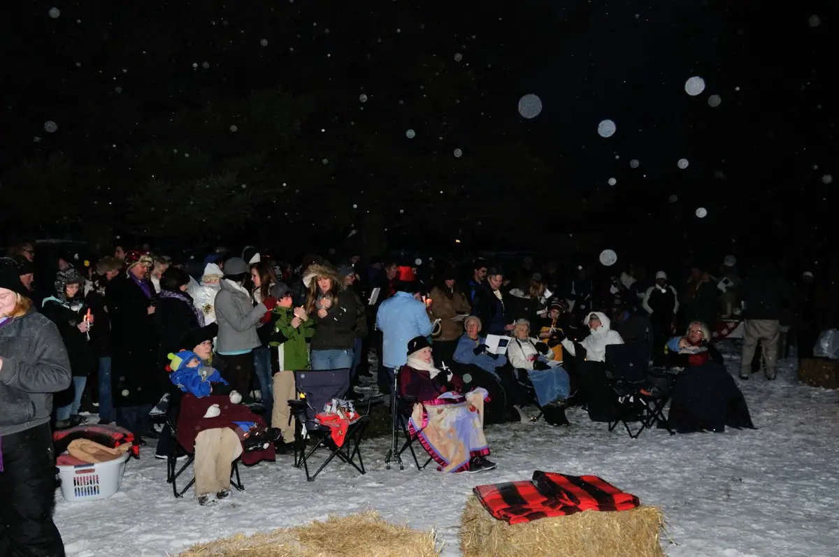 A bundled crowd gathers outdoors at night among folding chairs, candles, snow, and hay bales.