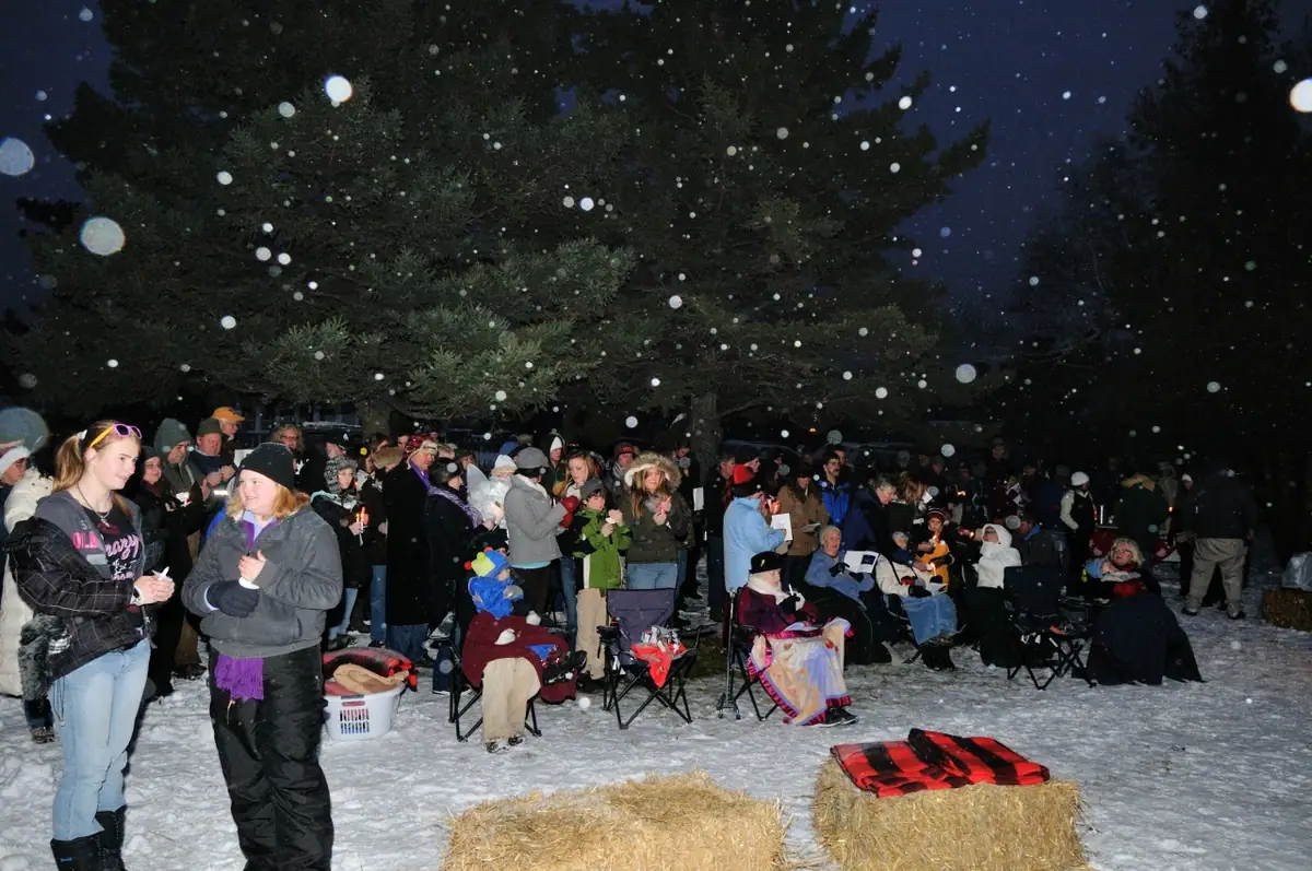 A large crowd gathers outdoors in falling snow among pine trees, folding chairs, candles, and hay bales.