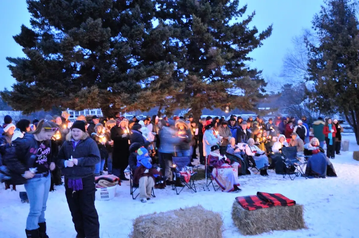 A candlelit gathering of people stands and sits in a snowy outdoor setting at dusk.