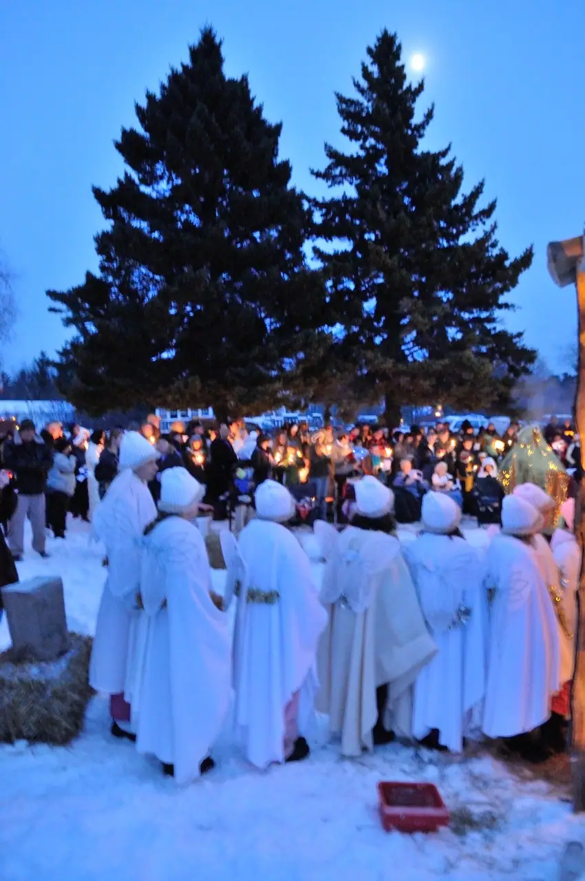 People in white robes gather in snow before a candlelit crowd beneath evergreen trees.