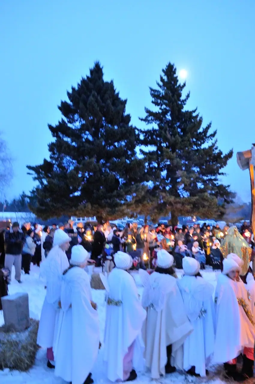 People in white robes gather in snow before evergreen trees as a candlelit crowd watches.