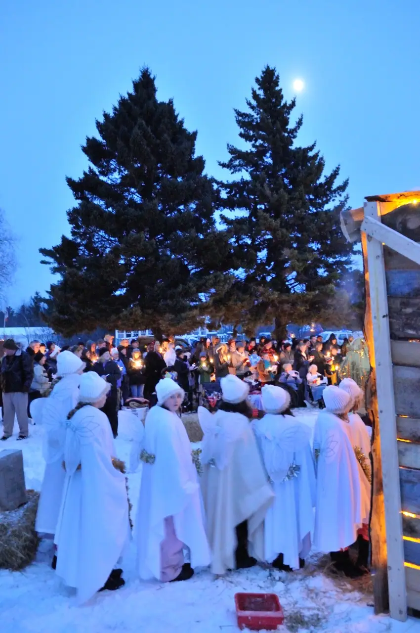 People in white angel costumes gather before a candlelit crowd beside a wooden structure in the snow.