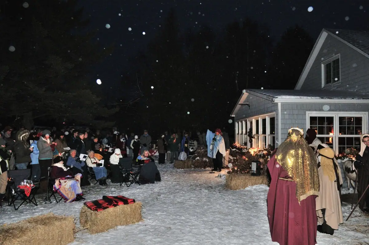 A nighttime outdoor gathering in snow, with costumed figures, hay bales, spectators, and a lit building.