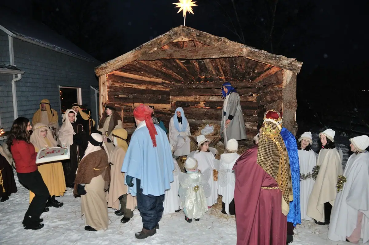 Costumed people gather around a rustic manger in a snowy nighttime nativity scene.