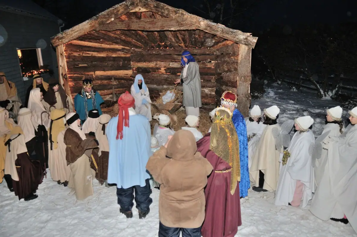People in costumes gather around a rustic wooden shelter in a snowy outdoor nativity scene at night.