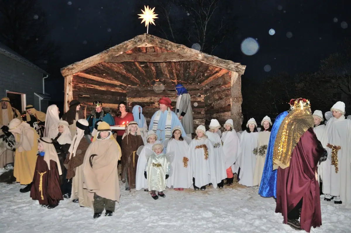 A costumed group gathers in front of a wooden shelter for an outdoor nativity scene in falling snow.
