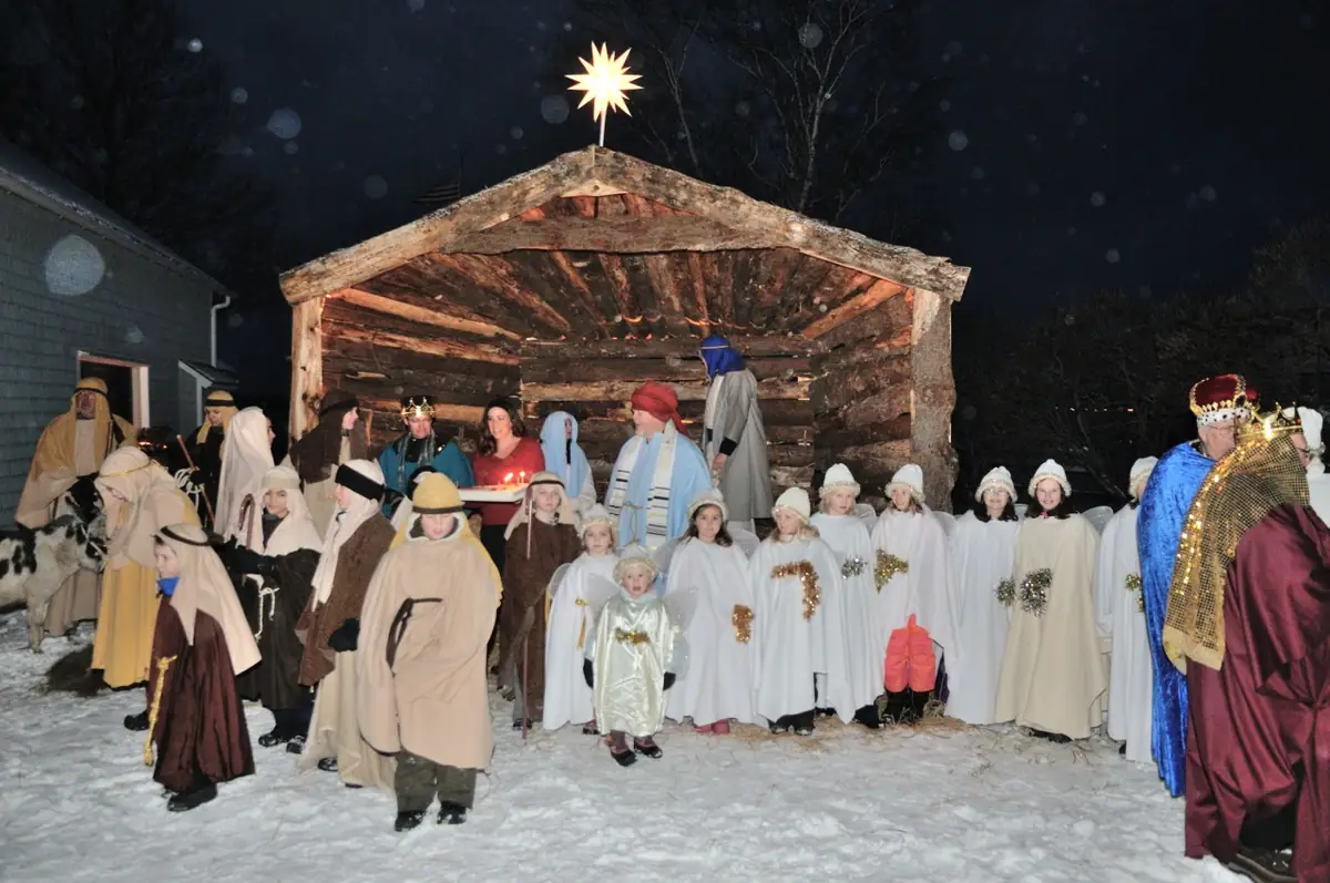 Costumed adults and children gather in the snow before a wooden nativity shelter topped by a lit star.