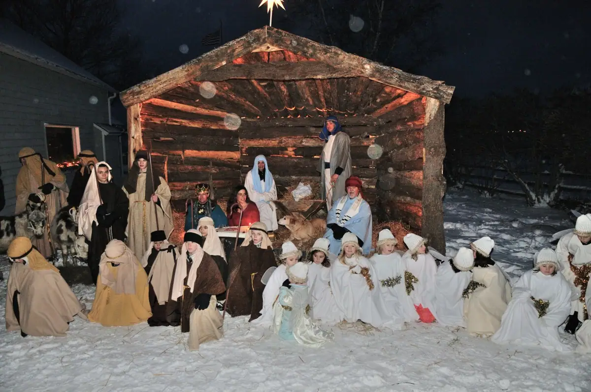 Costumed adults and children gather in the snow before a lit wooden stable beneath a star.