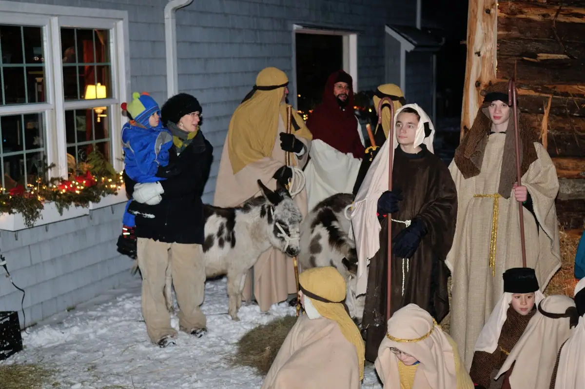 Costumed people and children gather with two goats outside a decorated building in the snow at night.