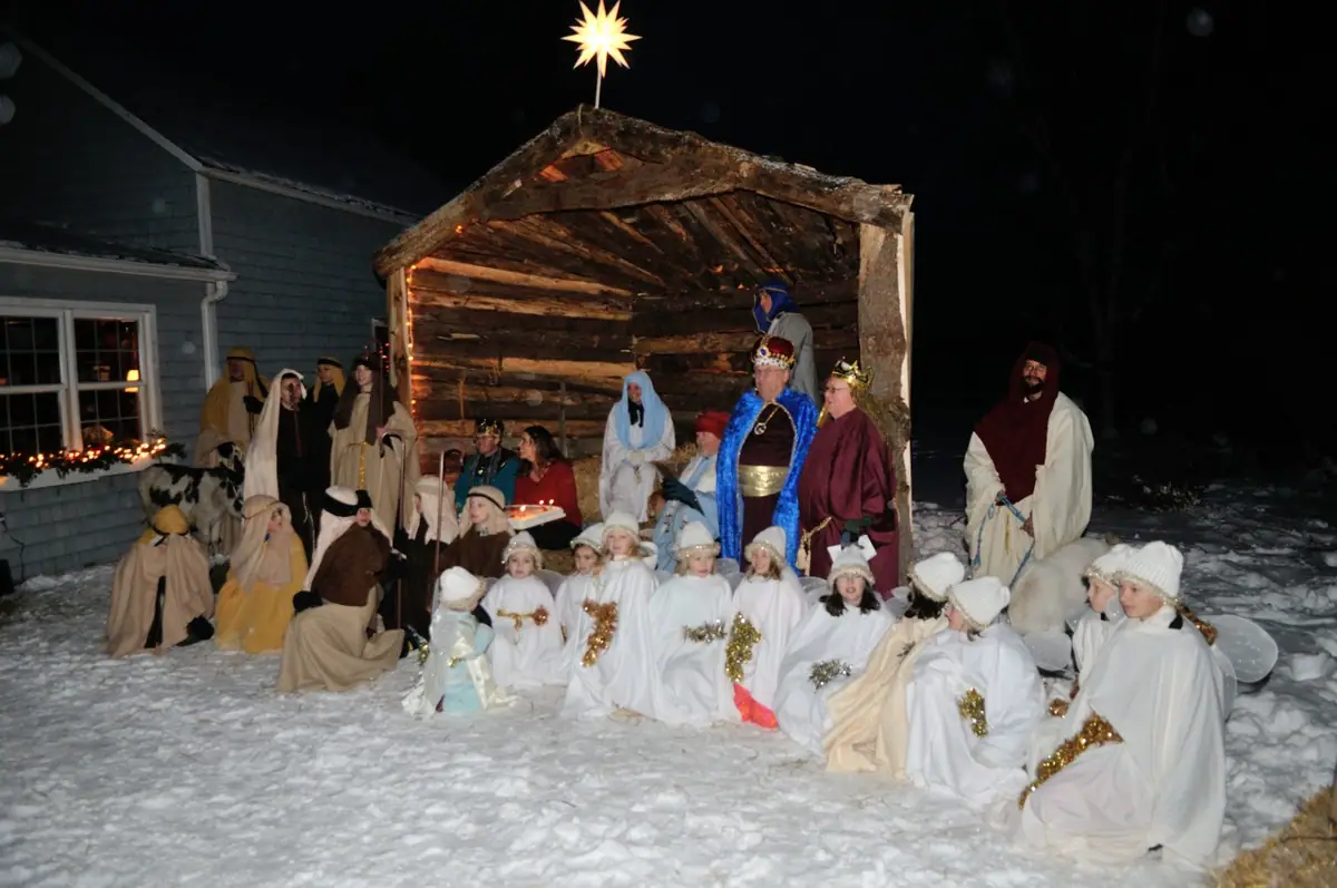 Costumed adults and children pose in a snowy outdoor nativity scene beside a rustic stable at night.