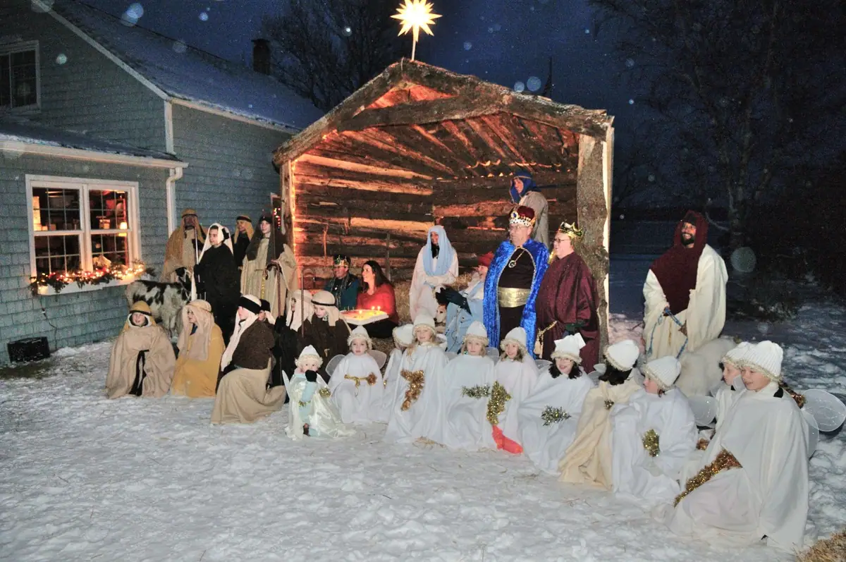 Costumed adults and children pose in the snow beside a lit wooden stable and a house at night.