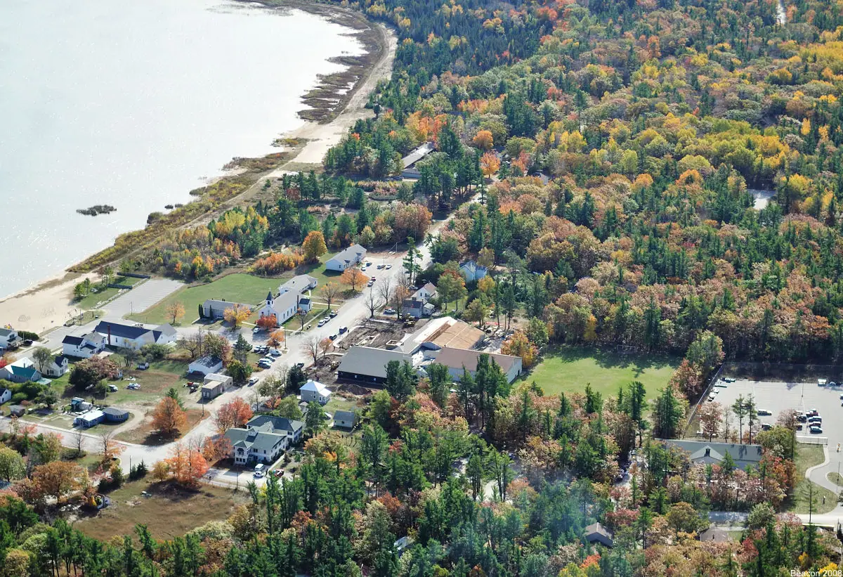 Aerial view of a wooded settlement with a shoreline, roads, homes, and a large institutional building.