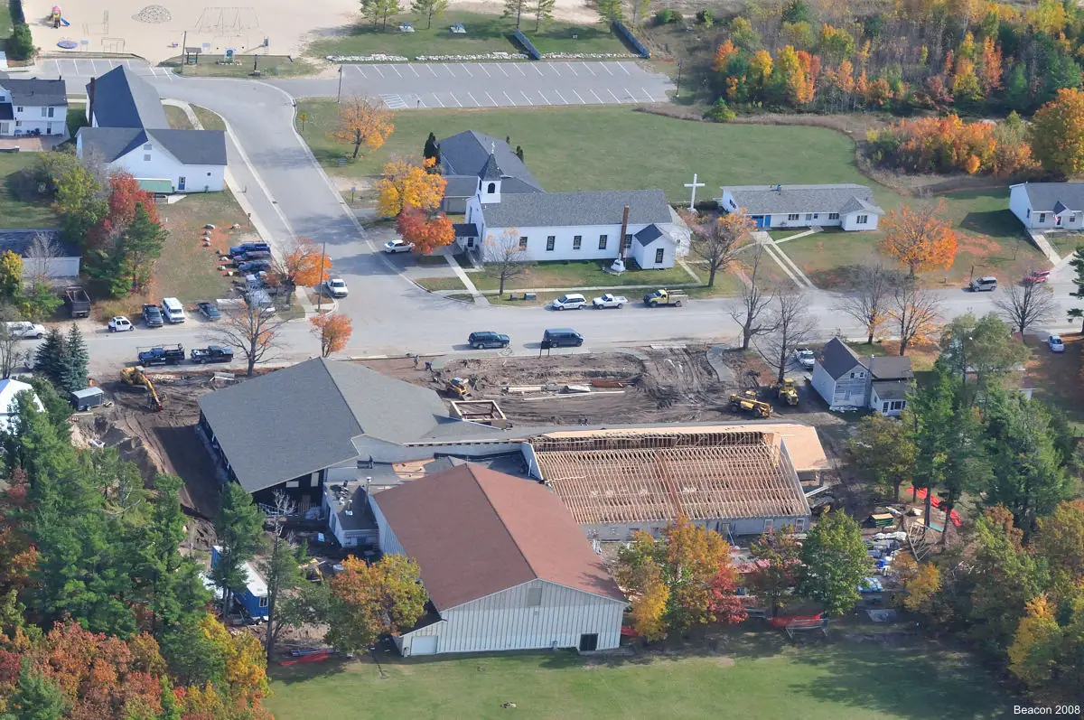 Aerial view of a school construction site, church, roads, vehicles, and autumn trees.