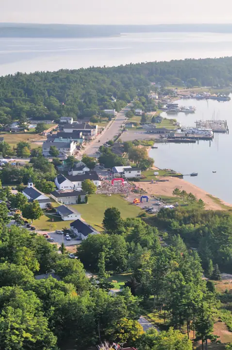 Aerial view of a waterfront community with docks, boats, roads, wooded areas, and a colorful gathering near the shore.