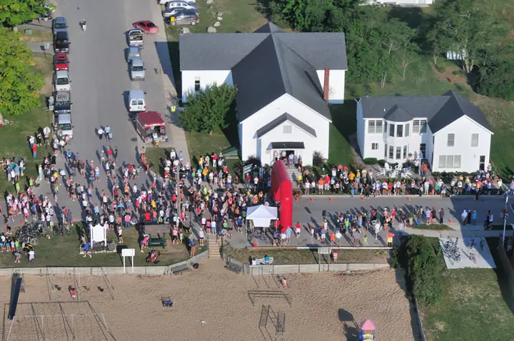 Aerial view of a road race gathering with runners, spectators, houses, vehicles, and a red inflatable arch beside a sandy beach.