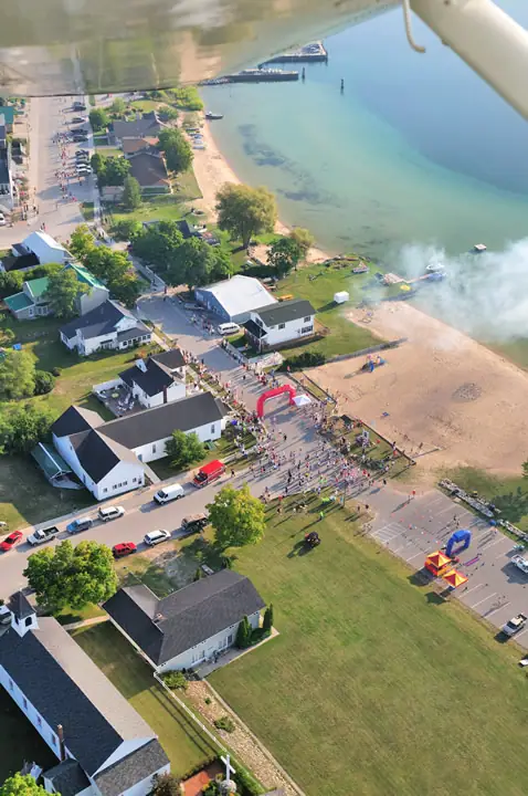 Aerial view of a shoreline race gathering with a start arch, homes, roads, and a pier.