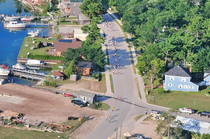 Aerial view of runners on a road beside docks, boats, homes, trees, and a construction lot.