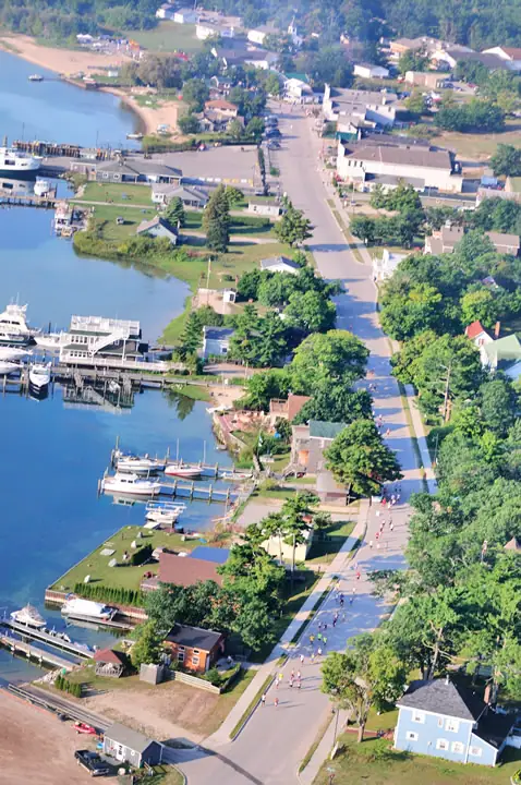 Aerial view of runners on a waterfront road beside docks, boats, homes, and tree-lined streets.