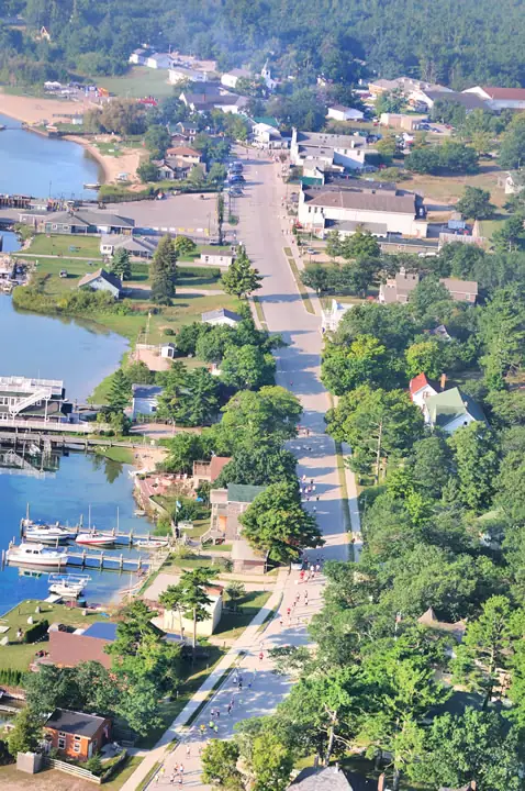 Aerial view of a waterfront neighborhood with boats, docks, a broad road, houses, and people along the roadway.
