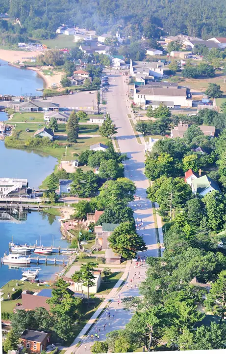Aerial view of a tree-lined waterfront street with boats, buildings, docks, and people along the roadway.