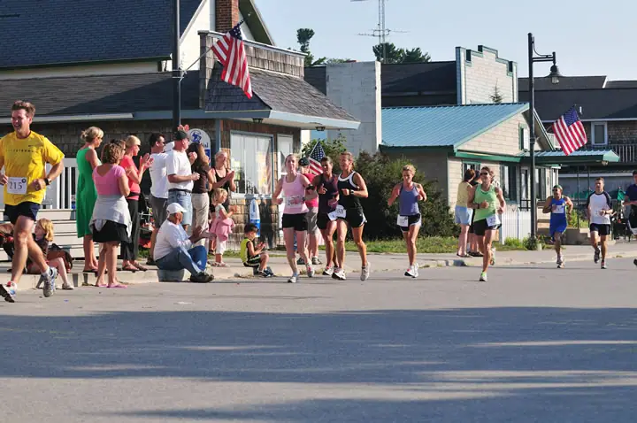 Runners pass spectators on a paved street lined with buildings and American flags.