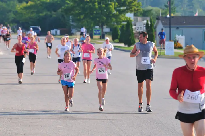 Runners wearing numbered bibs move along a paved road, with houses, trees, and a speed-limit sign behind them.