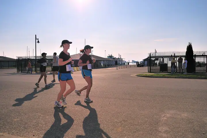 Runners cross a paved waterfront area as sailboat masts and spectators appear behind them.
