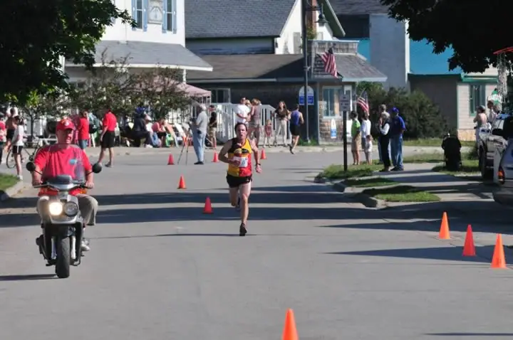 A runner and scooter rider move along a coned-off street while spectators watch from the sidewalks.