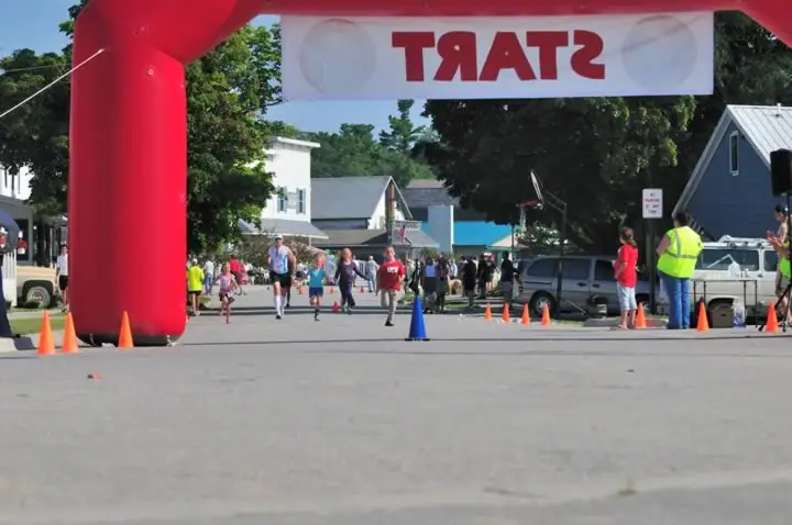 Runners begin a road race beneath a red inflatable arch, with cones and spectators lining the street.