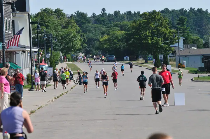 Runners and spectators fill a broad paved street beneath trees, with flags, bicycles, and roadside buildings visible.
