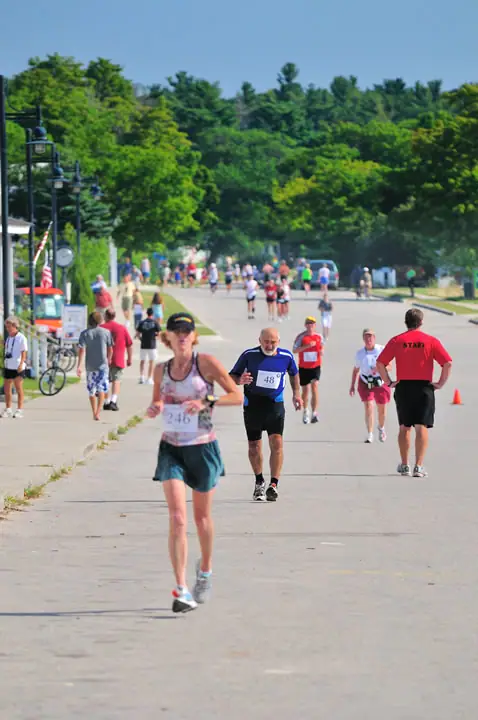 Runners and spectators fill a tree-lined paved road during a road race.
